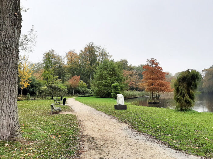 Caminho ao longo do lago no Amstelpark, com bancos, obras de arte e muitas árvores.